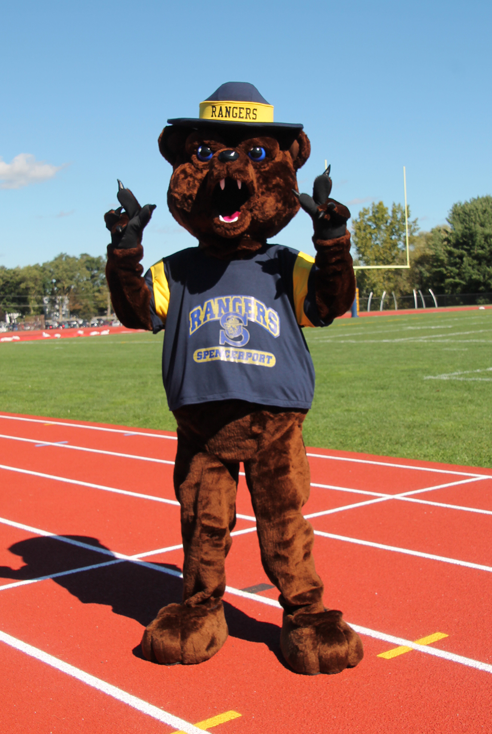 A fluffy brown bear mascot wearing a blue and yellow shirt that says "Rangers Spencerport" stands on a red running track.
